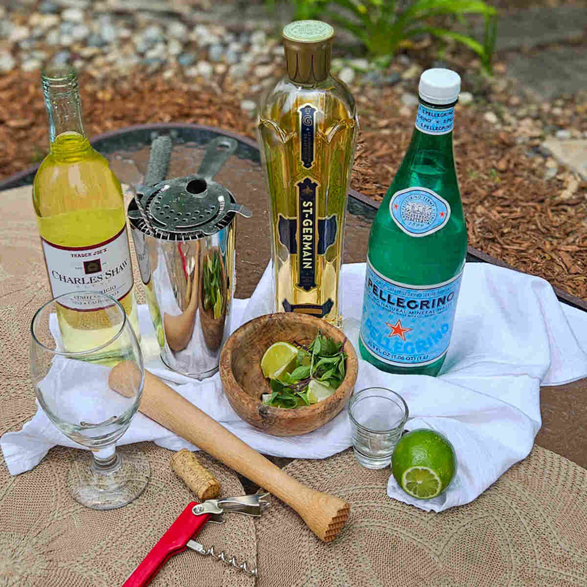 A table with a bottle of wine, a bottle of water, a bottle of St Germain, a bowl with limes and Thai basil. A muddler, wine opener, a cocktail shaker, and a shot glass.