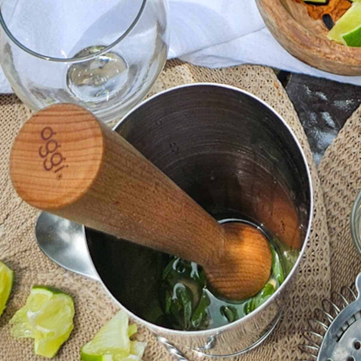 A wooden muddler is in a metal cup with limes and other ingredients.