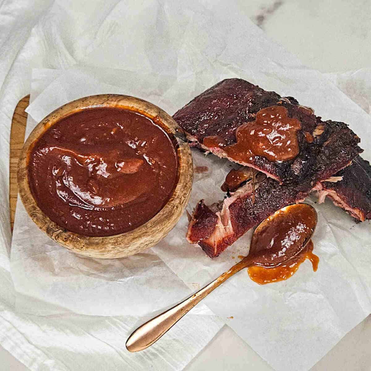 Barbecued ribs with a spoonful of sauce on top, a spoon of sauce and a bowl of it.