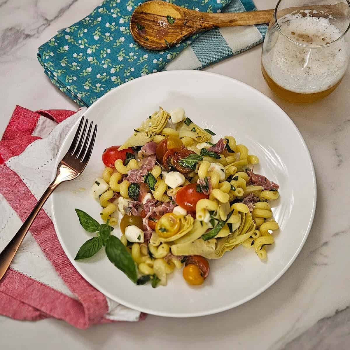 pasta salad with tomatoes, prosciutto, and fresh mozzarella on a plate with a fork and a beer.