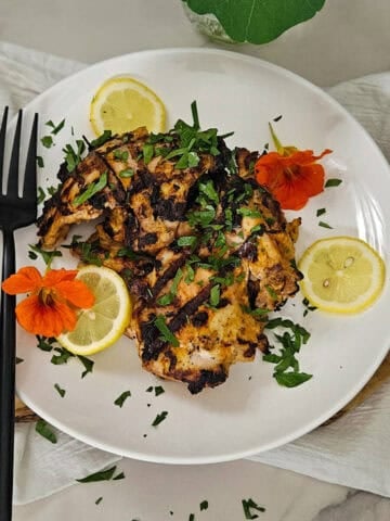 Grilled Greek yogurt marinated chicken with lemon slices and fresh herbs, served on a white plate.