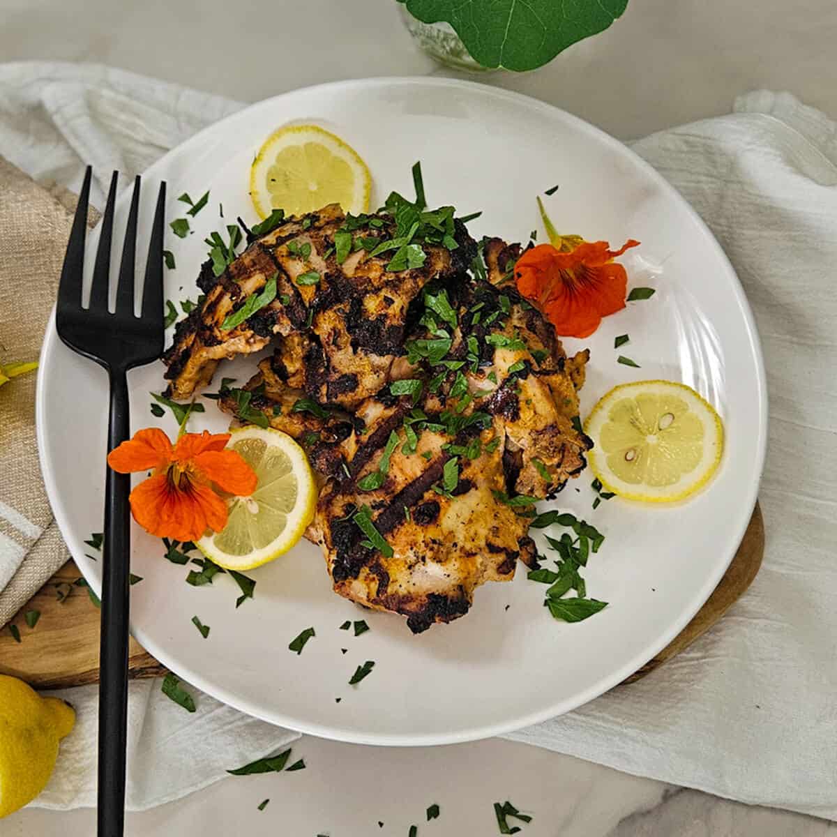 Grilled chicken thighs on a plate garnished with chopped fresh parsley, lemon, flowers and a fork
