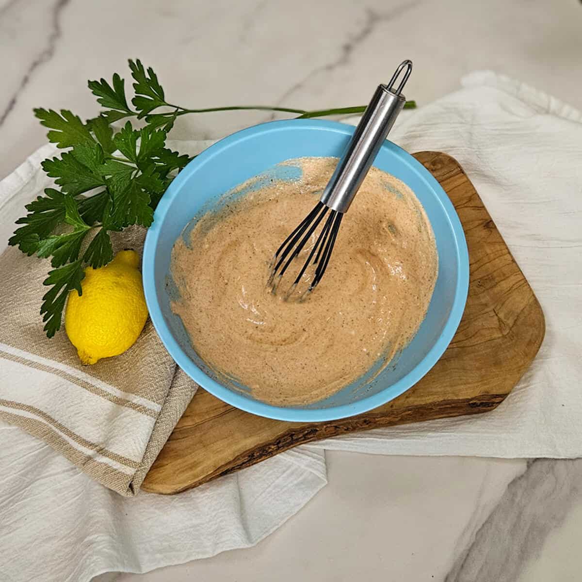 Creamy Greek yogurt marinade being whisked in a blue bowl on a wooden board. Fresh parsley and lemon garnish for flavor enhancement.
