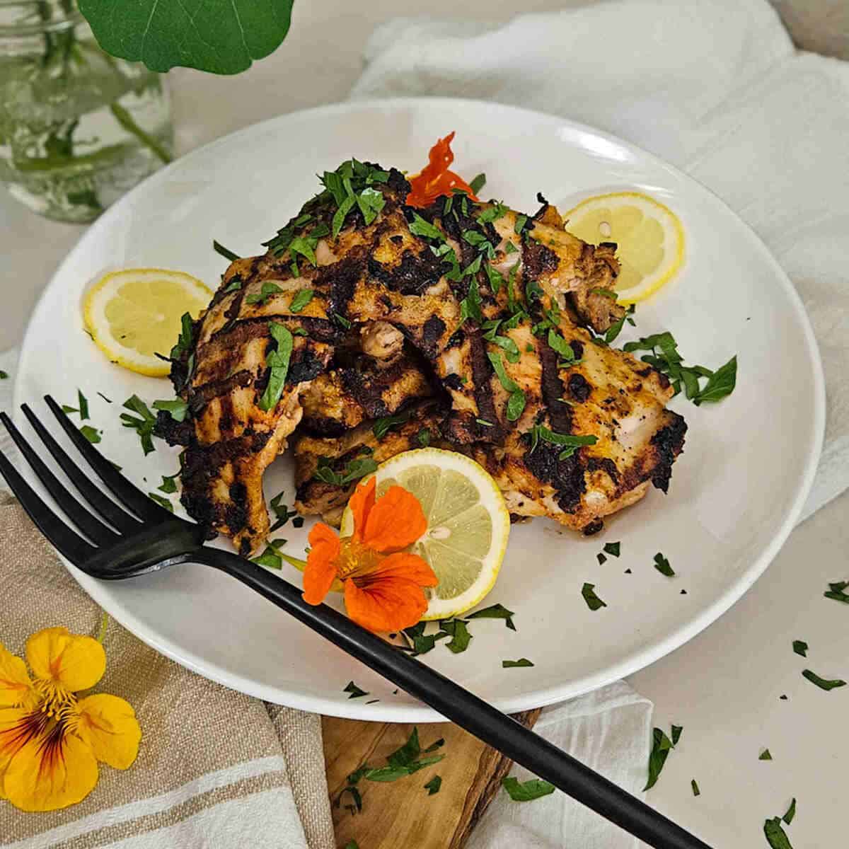 Grilled Greek yogurt marinated chicken seasoned with herbs on a white plate with lemon slices and fresh herbs, perfect for Season Two Taste.