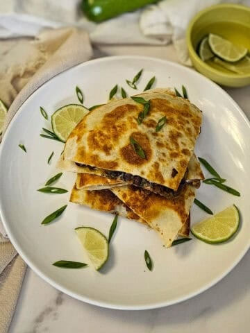 Golden-brown quesadilla with black beans and melted cheese, garnished with lime wedges and fresh herbs.