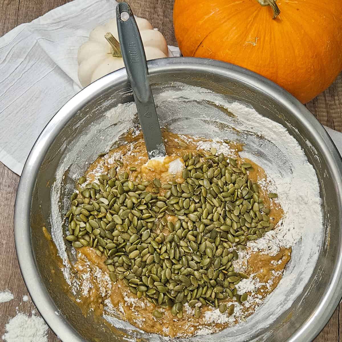 Adding pumpkin seeds to the zucchini pumpkin spice bread batter in mixing bowl with pumpkin, flour, and seeds on top.