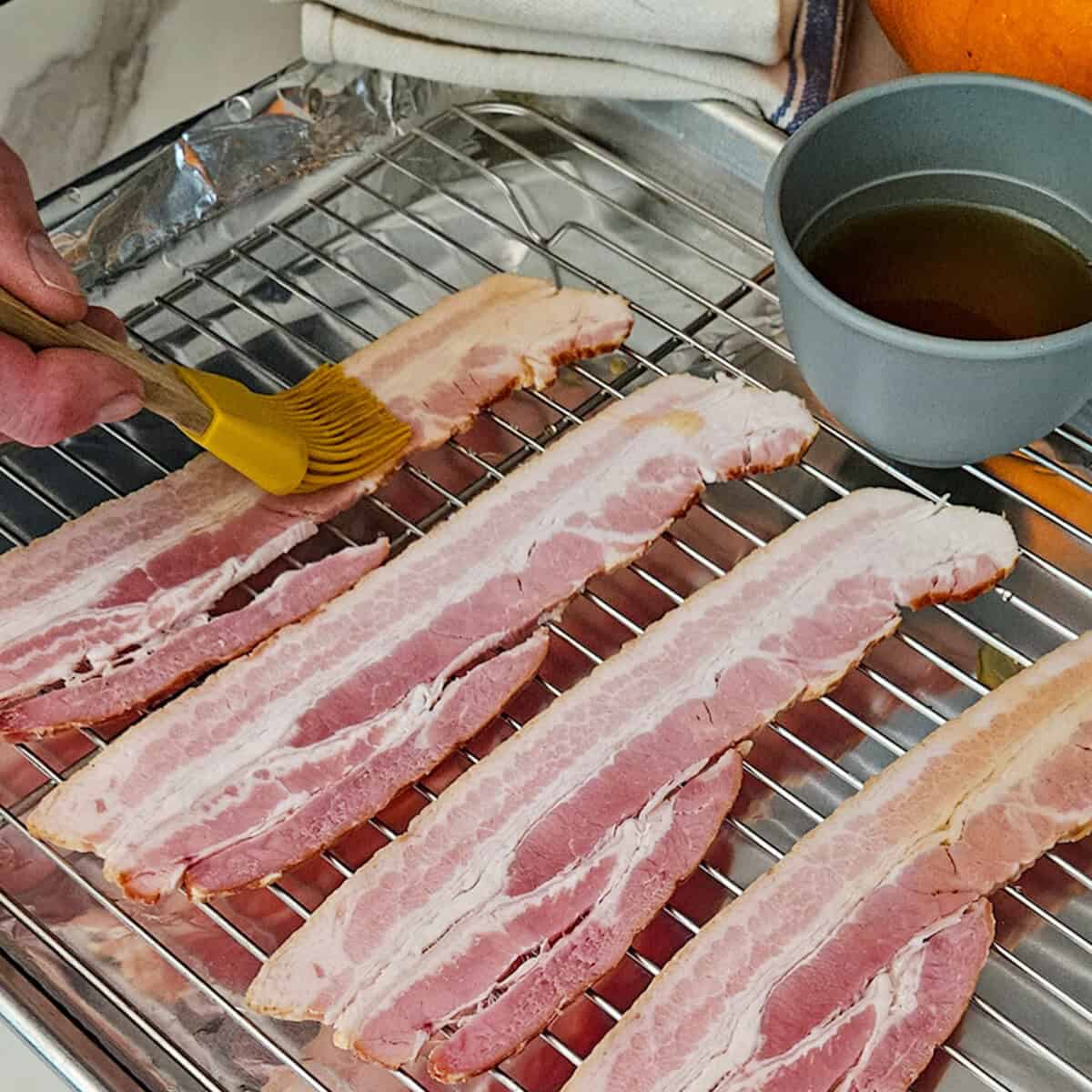 Bacon strips being brushed with maple syrup on a wire rack, ready for cooking or seasoning.