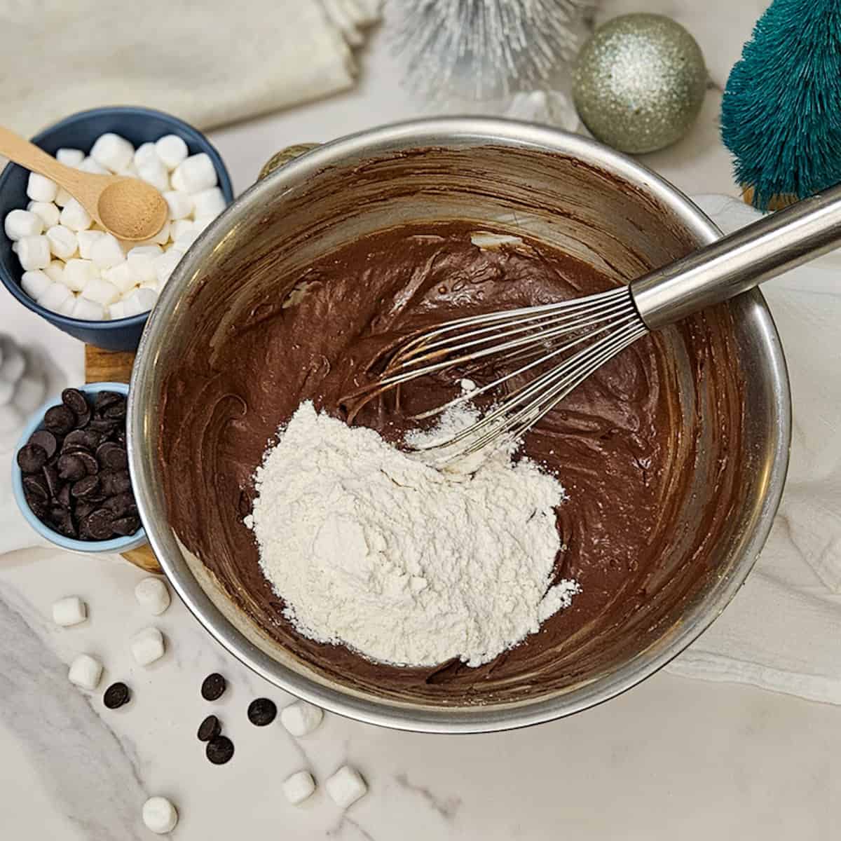 Adding flour to creamy chocolate batter with marshmallows, mini chocolate chips, and holiday decorations.