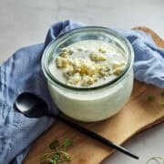 Blue cheese dressing in a glass jar with crumbles on top, served on a wooden board with a blue cloth.