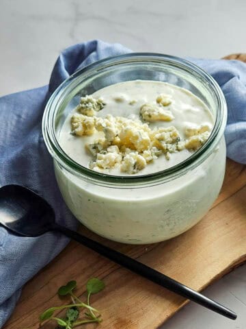 Blue cheese dressing in a glass jar with crumbles on top, served on a wooden board with a blue cloth.