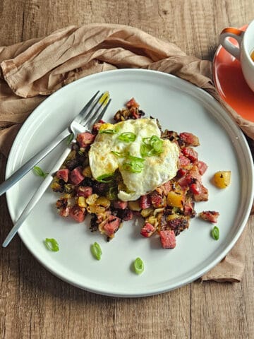 Corned beef hash on a white plate with a fried egg and sliced green onions and a fork and knife, served with a cup of tea.