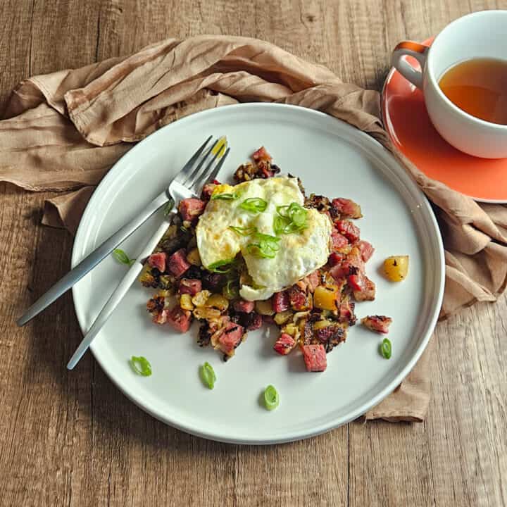 Corned beef hash on a white plate with a fried egg and sliced green onions and a fork and knife, served with a cup of tea.