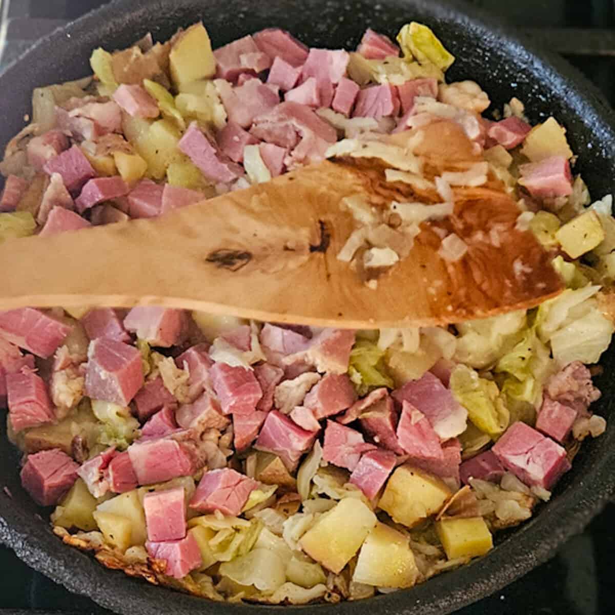 Stirring corned beef hash in a skillet with a wooden spatula.