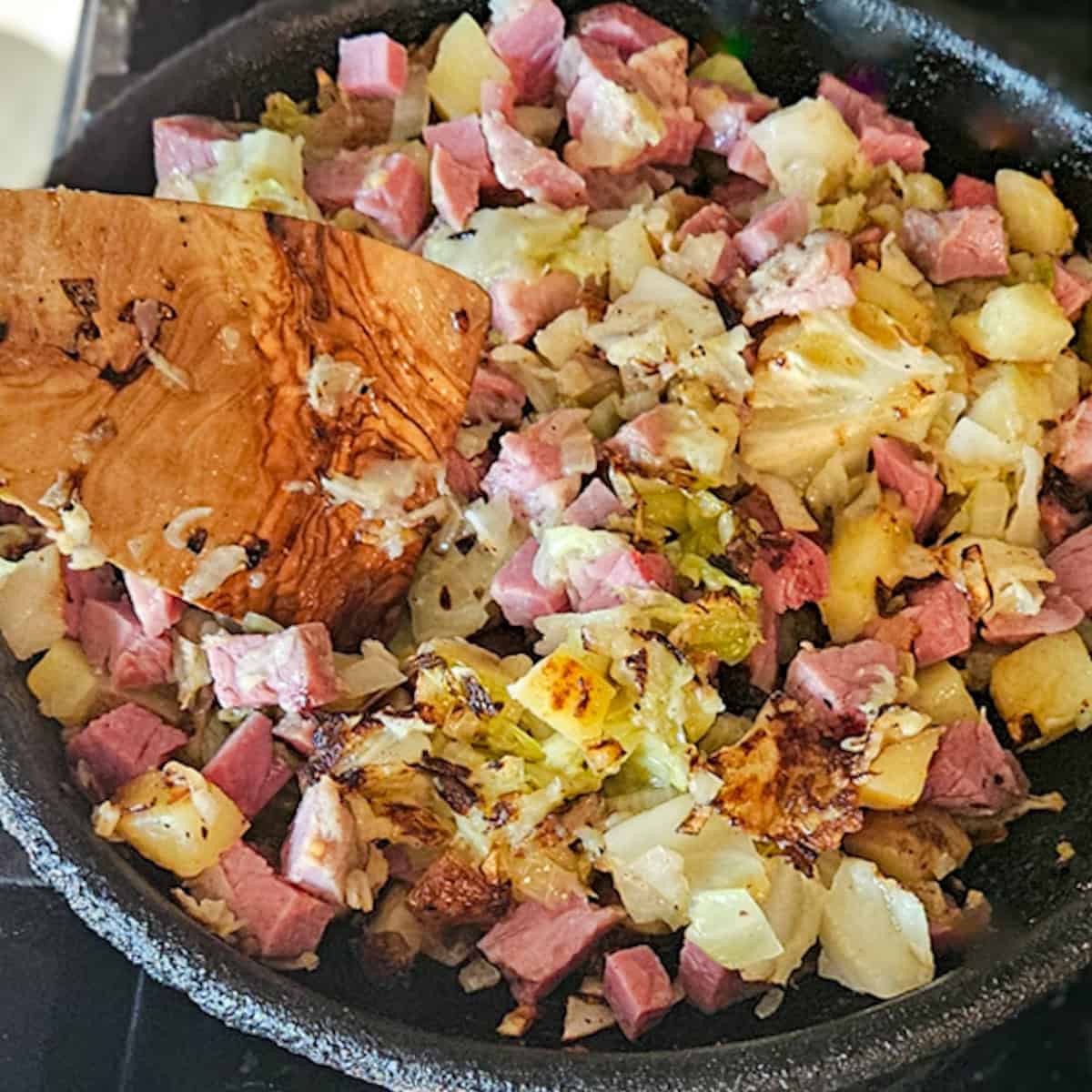 Stirring the cooked corned beef hash in a skillet with a wooden spatula.