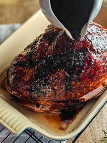 Pouring brown sugar balsamic glaze over the top of a ham in a baking dish.