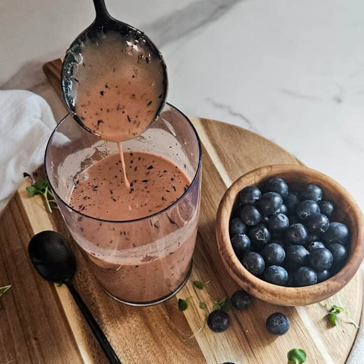 Blueberry balsamic dressing in a mixing cup with a spoon being lifted out of the cup, and a spoon, and a bowl of fresh blueberries.