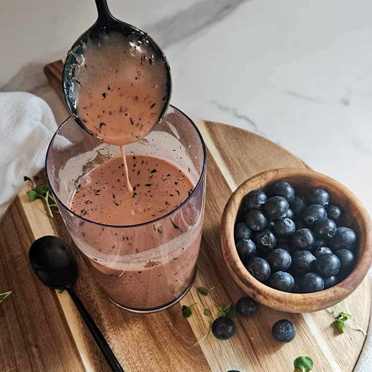 Blueberry balsamic dressing in a mixing cup with a spoon being lifted out of the cup, and a spoon, and a bowl of fresh blueberries.