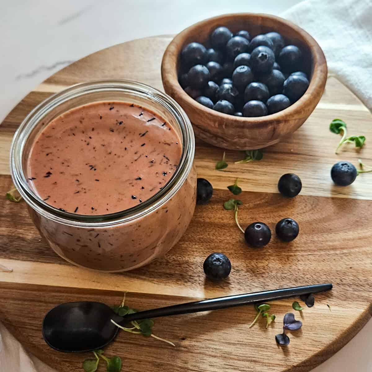 Blueberry balsamic dressing in a jar on a wooden board with fresh blueberries in a bowl and scattered on the board and a spoon.