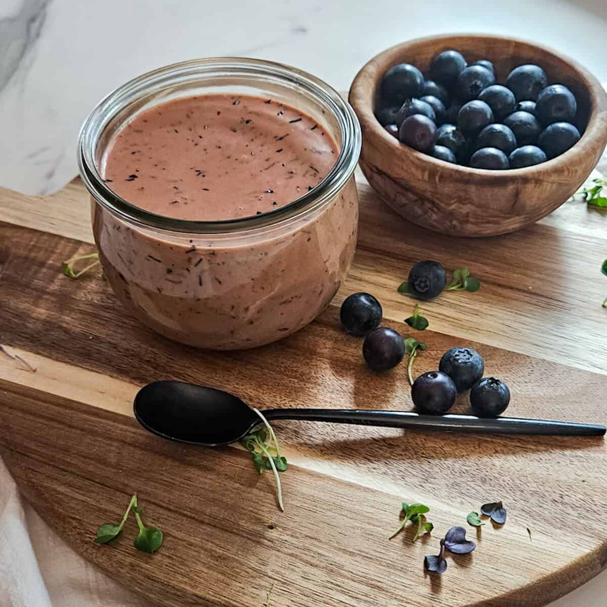 Blueberry balsamic dressing in a jar on a wooden board with fresh blueberries in a bowl and blueberries scattered on the board and a spoon.