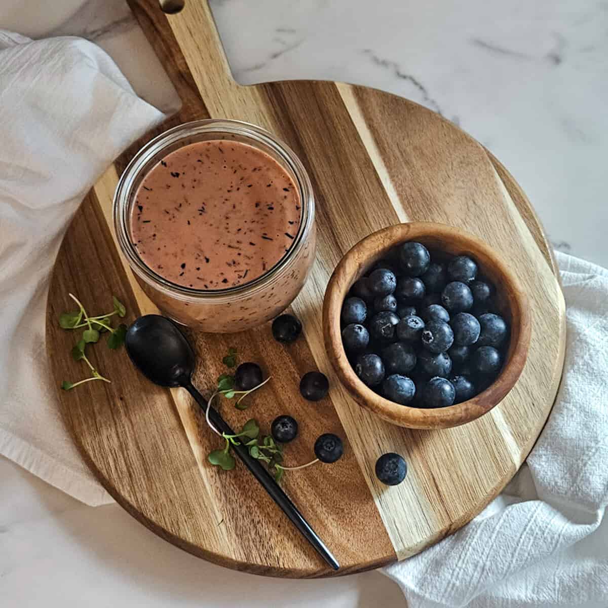 Blueberry balsamic dressing in a bowl on a wooden board with a bowl of fresh blueberries, a spoon, and blueberries and green sprouts scattered on the board.
