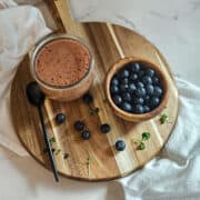 Blueberry balsamic dressing in a jar on a wooden board with a spoon, a bowl of fresh blueberries and blueberries and green sprouts scattered on the board.