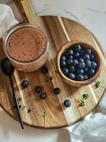 Blueberry balsamic dressing in a jar on a wooden board with a spoon, a bowl of fresh blueberries and blueberries and green sprouts scattered on the board.