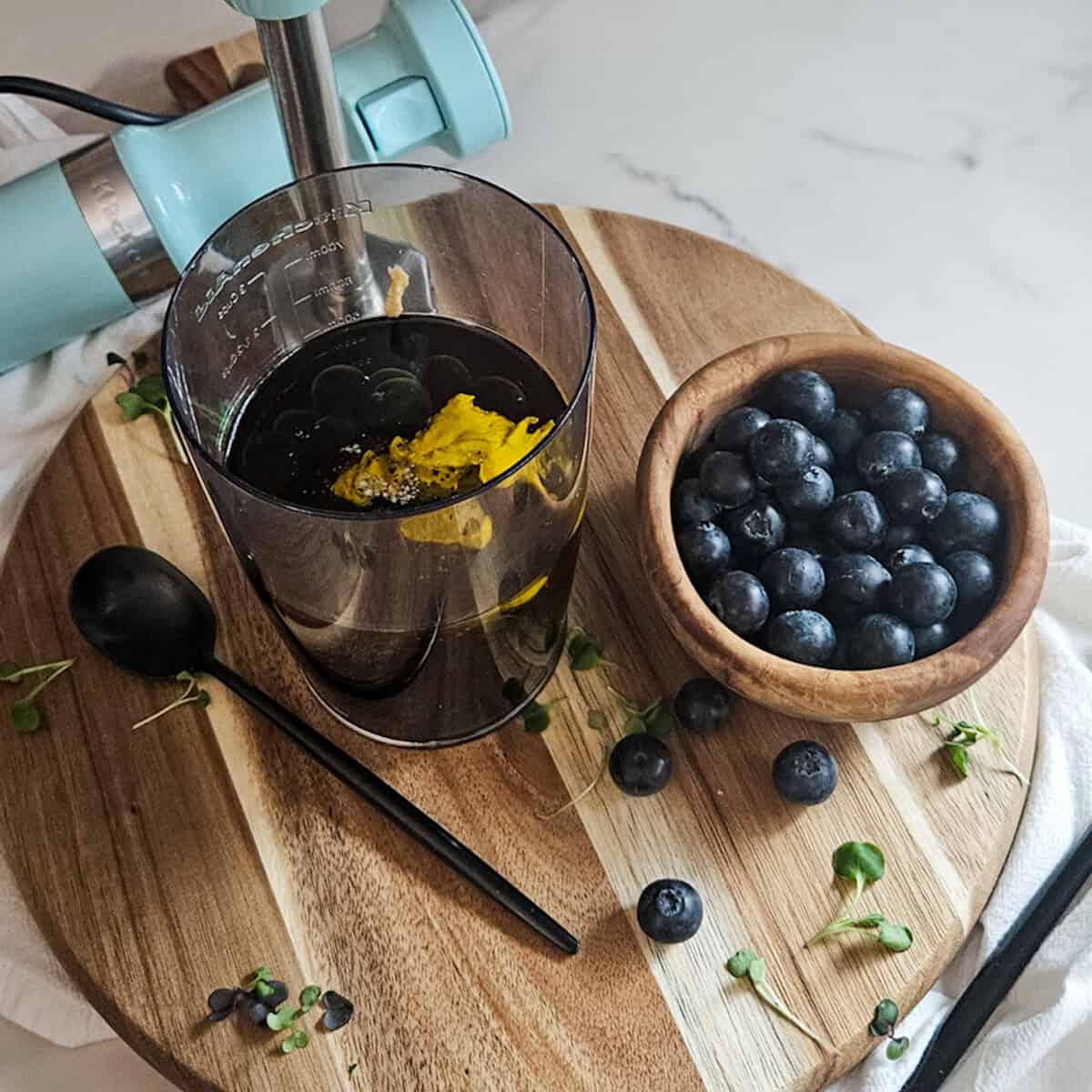 The ingredients for blueberry balsamic dressing in a mixing cup, a bowl of fresh blueberries, a spoon, and an immersion blender in the background.