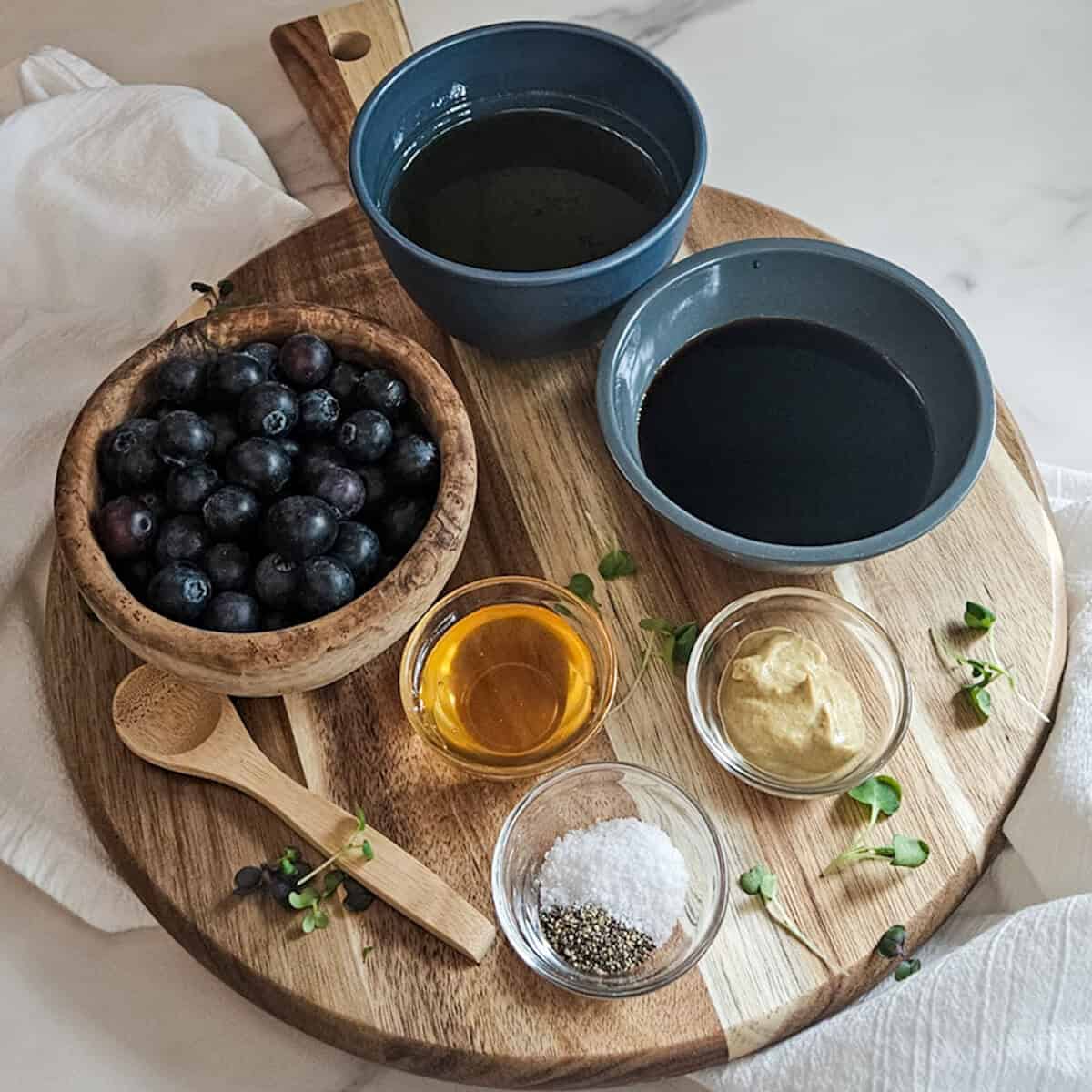 The ingredients for the blueberry balsamic dressing measured into separate bowls on a wooden board with a small spoon.