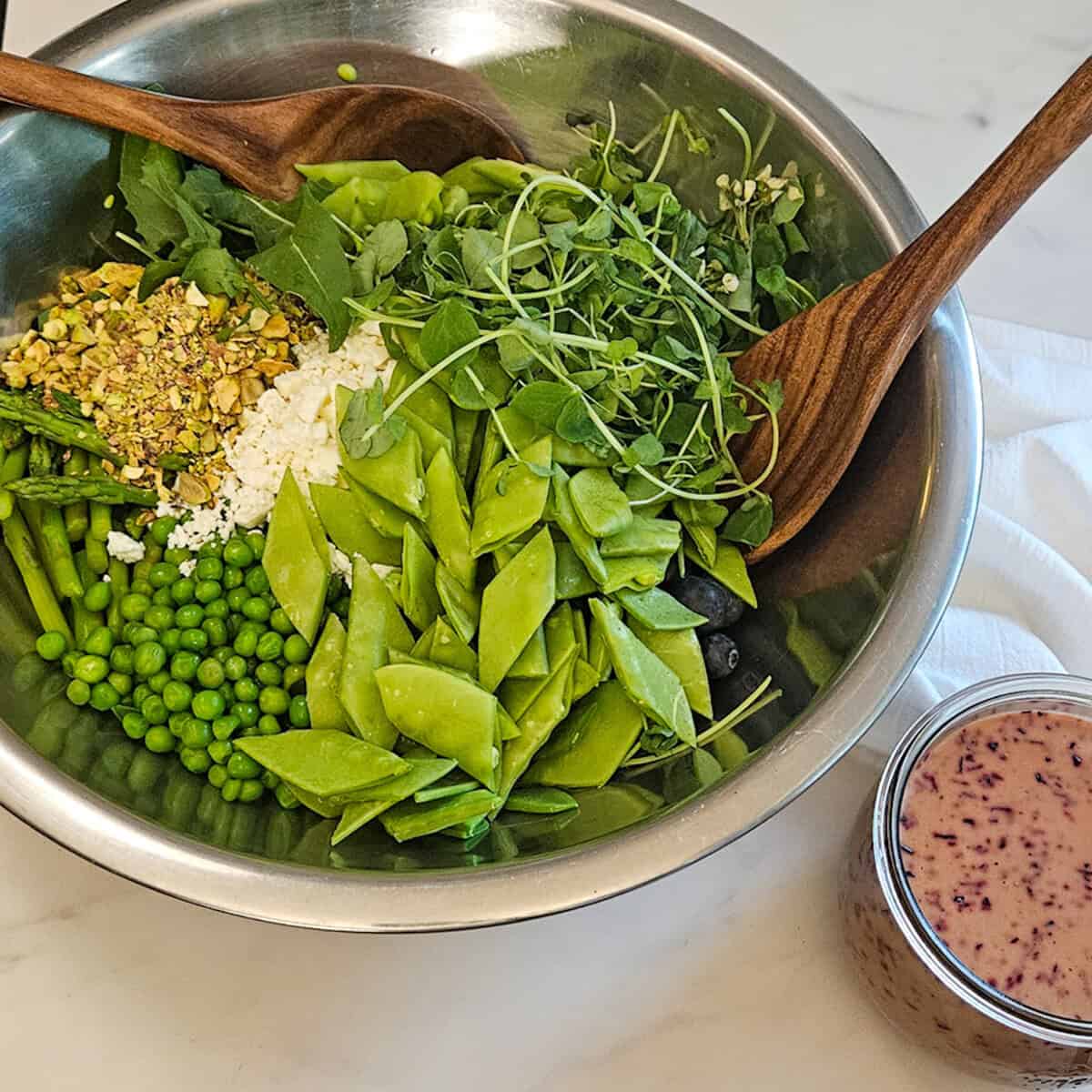 Mixing the ingredients for the pea and asparagus salad in a metal bowl with to wooden spoons and a jar of blueberry balsamic dressing along side.