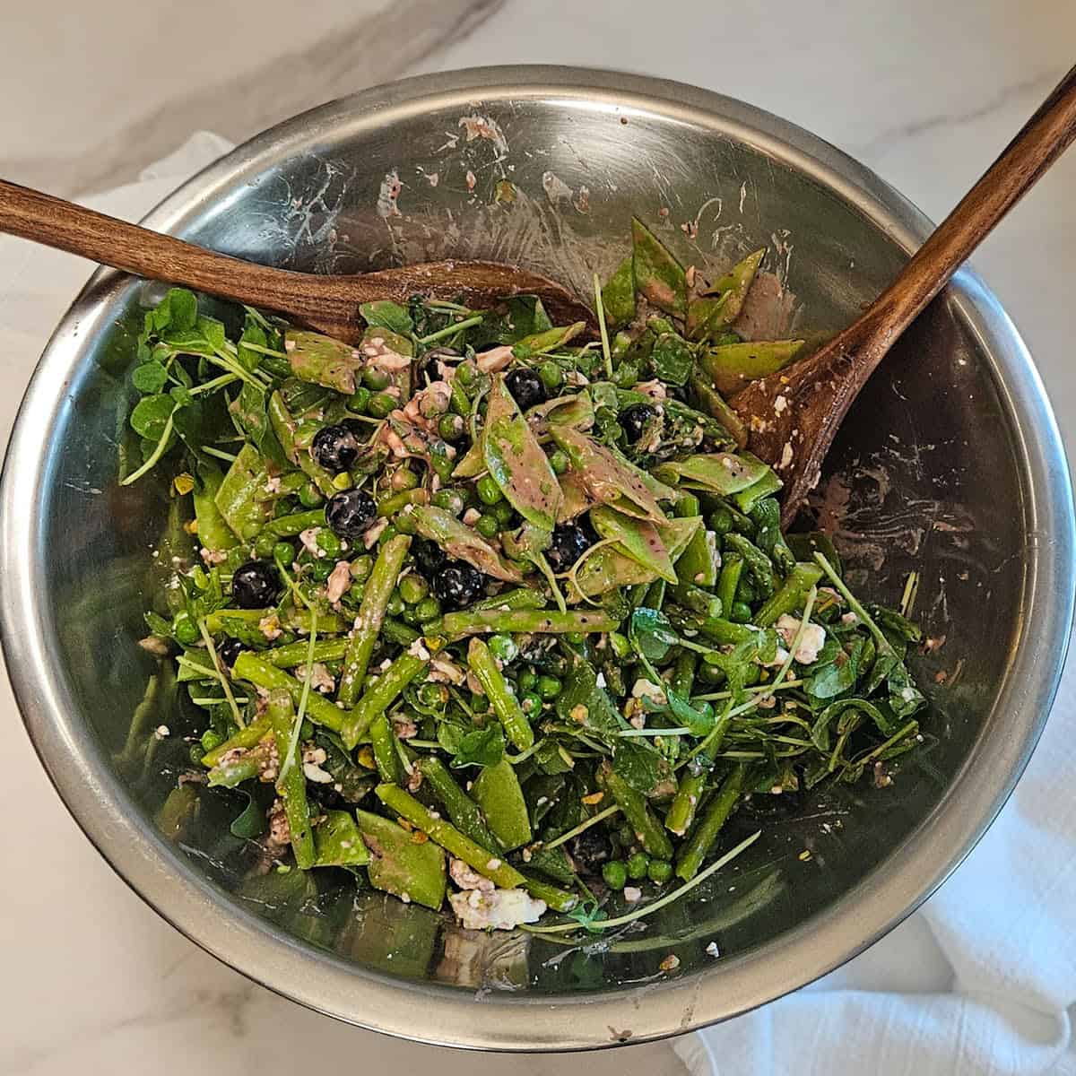 Mixing together the pea and asparagus salad in a metal bowl using two large wooden spoons.
