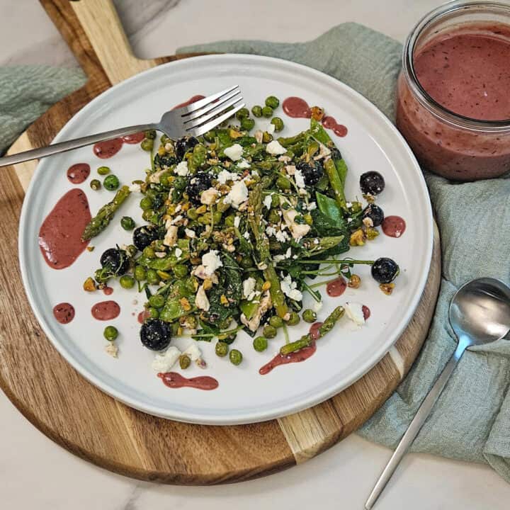 Pea and asparagus salad on a plate with a fork and spoon on a wooden board along side a jar of blueberry balsamic dressing.
