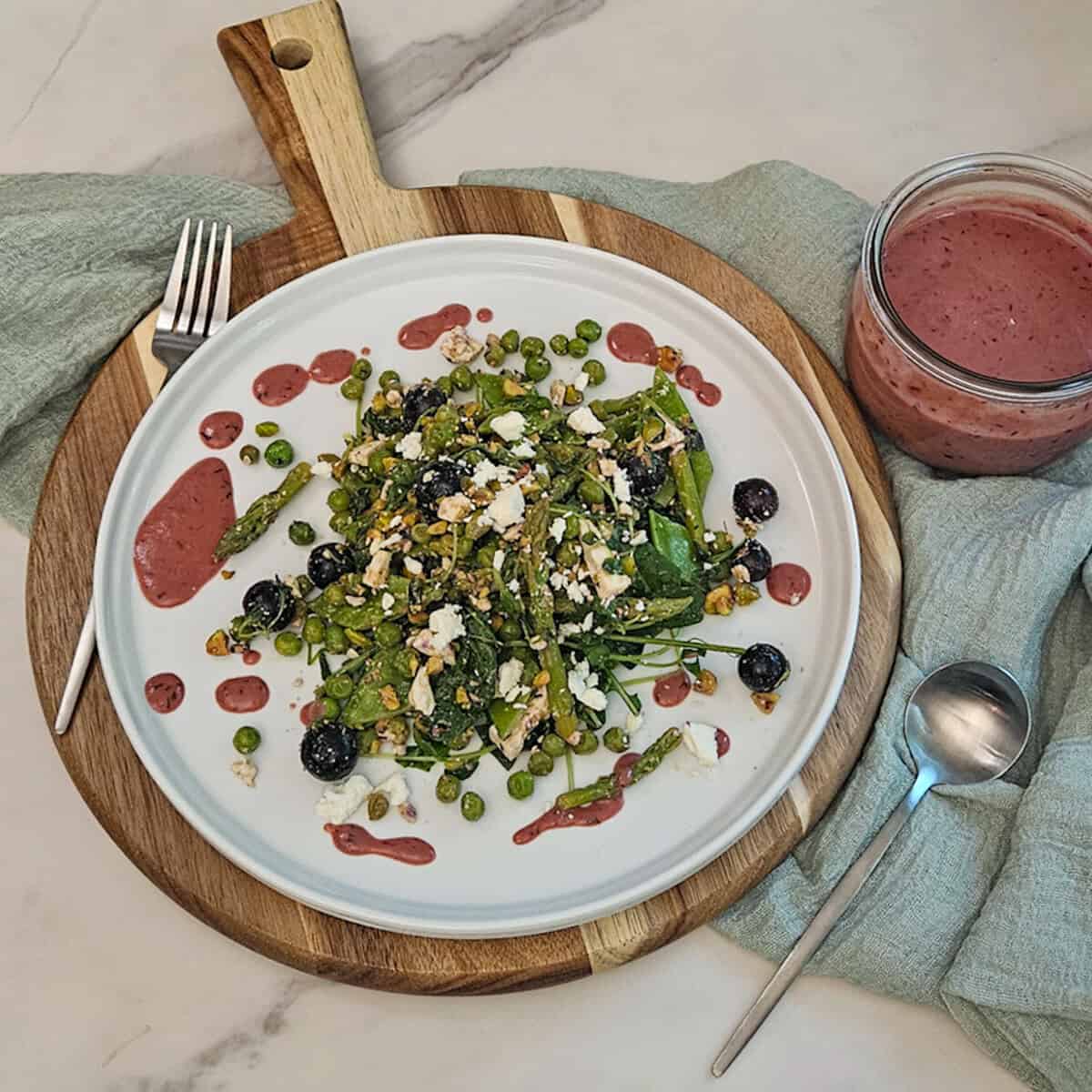 Pea and asparagus salad on a plate on a wooden board with a fork and spoon and along side a jar of blueberry balsamic dressing.