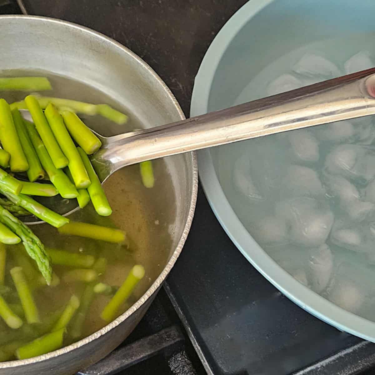 Using a large metal spoon to move asparagus from a pot with hot water to a bowl containing ice water.