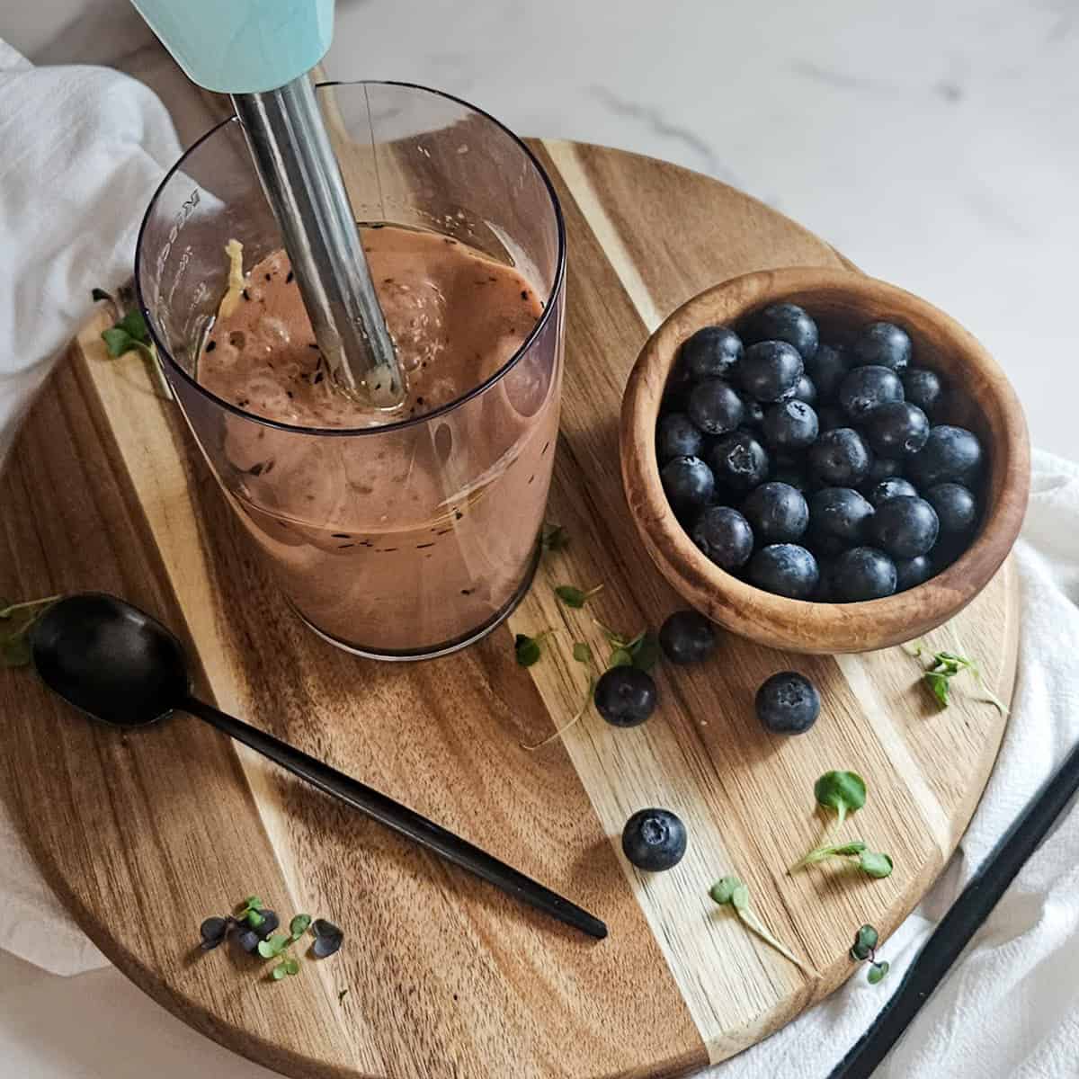 Mixing blueberry balsamic dressing in a mixing cup with an immersion blender on a wooden board with a spoon and a bowl of fresh blueberries.