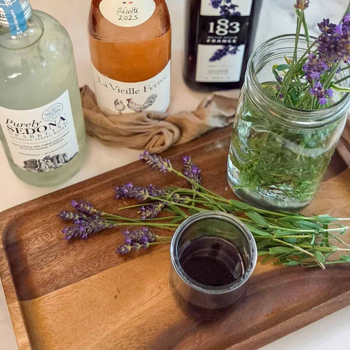 A bottle of sparkling water, lavender syrup, and rose` with fresh lavender flowers and a jar with lavender syrup on a wooden tray.