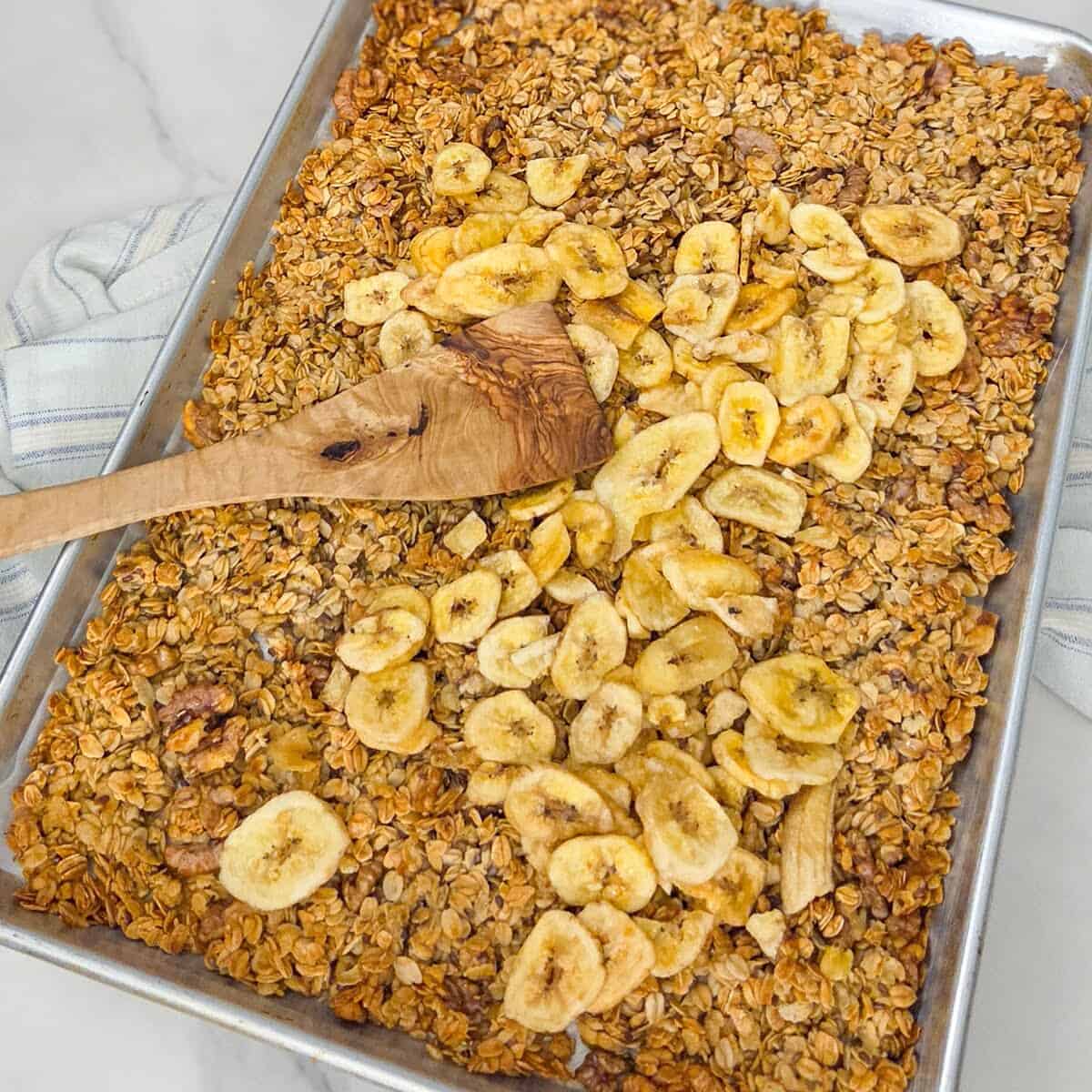 Cooked banana nut granola on a baking sheet pan with banana chips on top and a spatula.