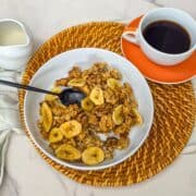 A bowl of banana walnut granola with a spoon , a cup of black coffee, and a pitcher of milk.