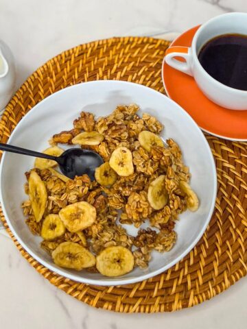 A bowl of banana walnut granola with a spoon , a cup of black coffee, and a pitcher of milk.