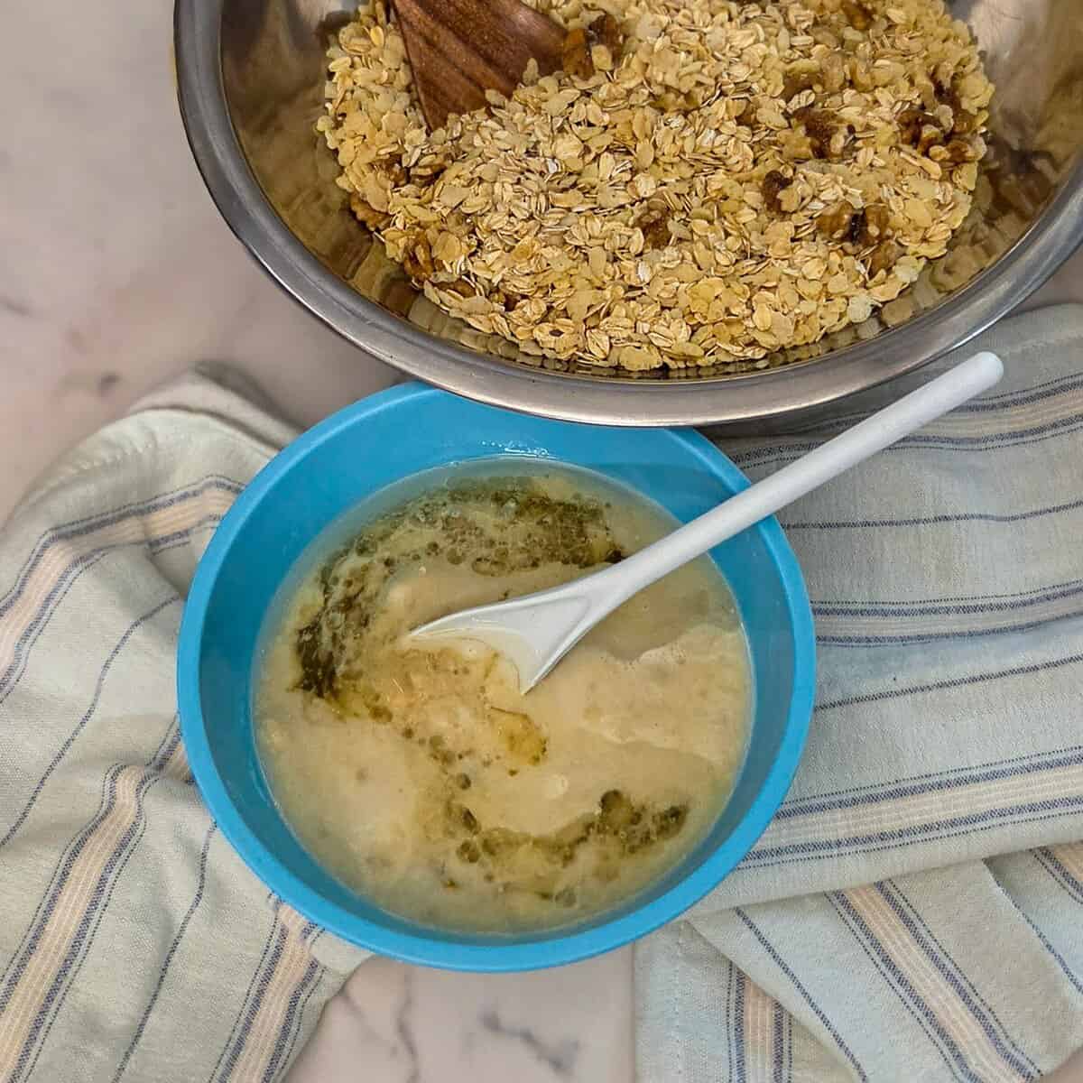 Two mixing bowls one containing the banana and oil mixture and the other with the cereal and oat mixture with spoons.