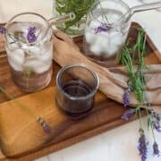 Two glasses with straws with lavender rose` spritzes on a wooden tray with lavender flowers and a jar of lavender syrup.
