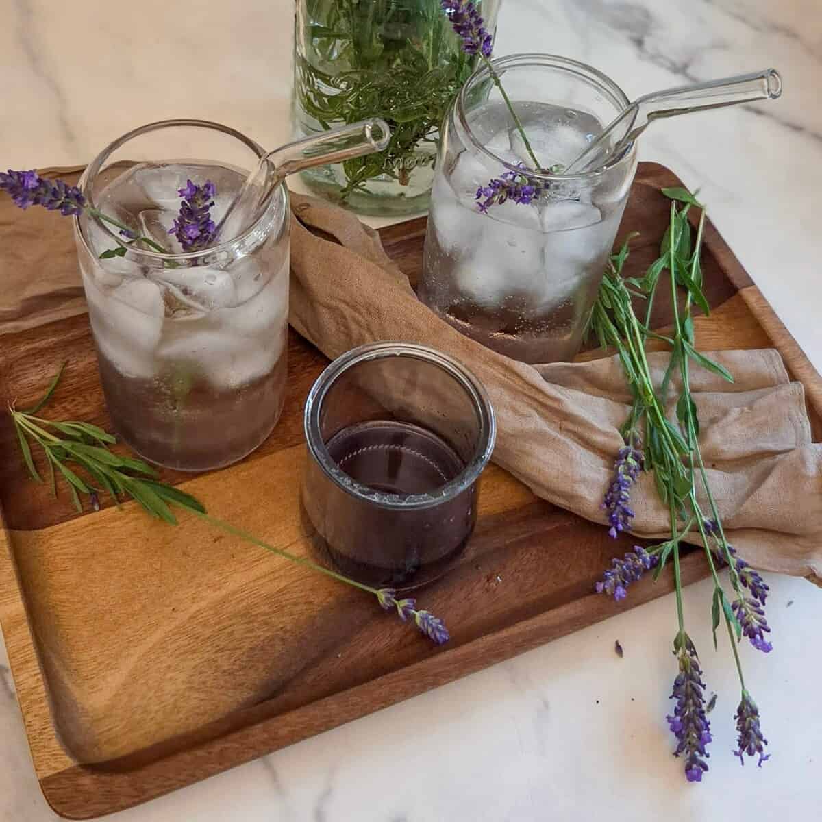 Two glasses with lavender rose` spritzes, lavender flowers, and a jar with lavender syrup on a wooden tray.