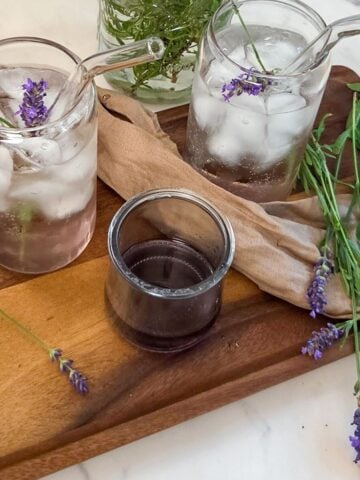 Two glasses with straws with lavender rose` spritzes on a wooden tray with lavender flowers and a jar of lavender syrup.