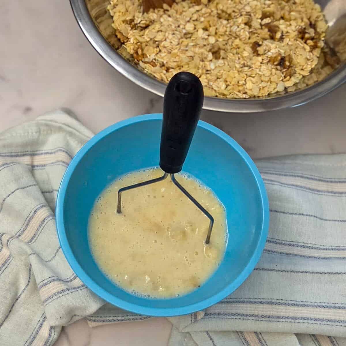 Using a potato masher to mash the sliced banana and oat milk in a blue bowl along side a bowl containing oats, walnuts, and cereal.