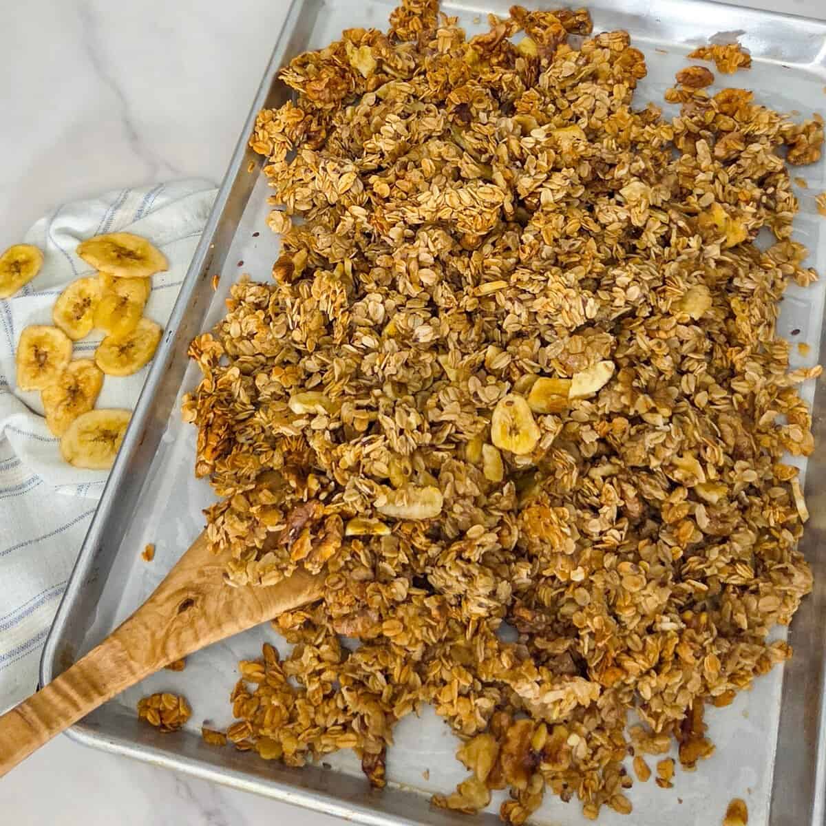 Mixing the banana chips into the cooled granola on a baking sheet pan with a spatula.