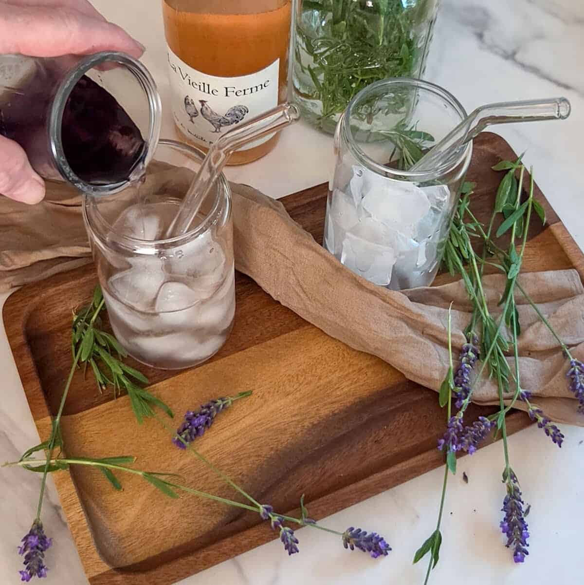 Pouring lavender syrup into a glass with rose`, ice, and a straw on a wooden tray with lavender flowers and a glass with ice and a straw, and a bottle of rose`.