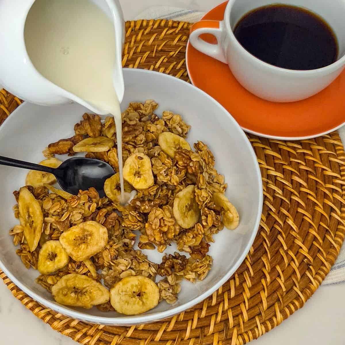 Pouring milk from a pitcher into a bowl of banana nut granola with a spoon along side a cup of black coffee.
