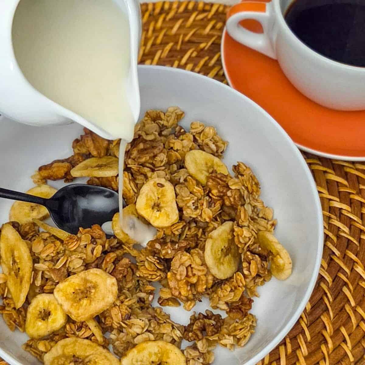 A closeup of pouring milk into a bowl of banana walnut granola with a spoon along side a cup of black coffee.