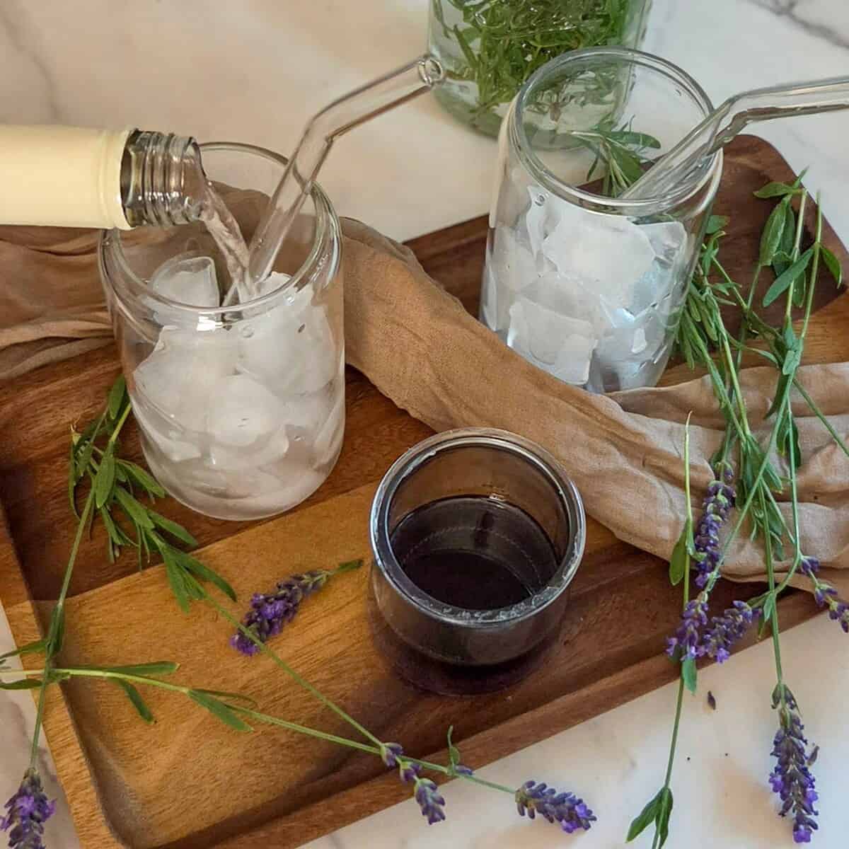 Pouring rose` into a glass with ice and a straw, on a tray with lavender flowers and a glass with ice and a straw.