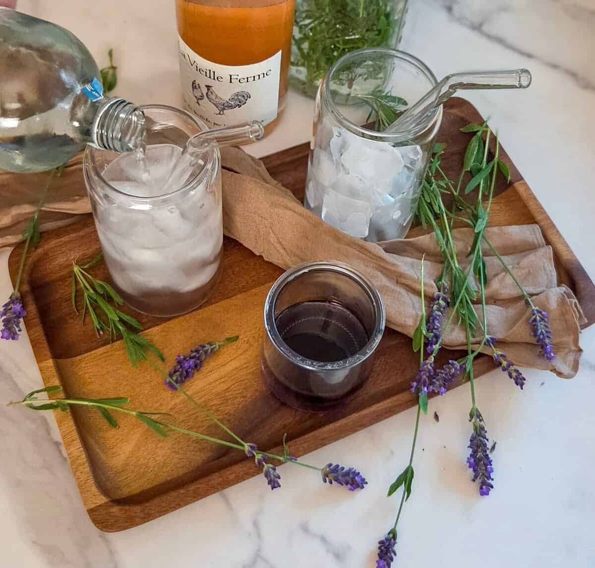 Pouring sparkling water into a glass with rose` and lavender syrup on a wooden tray with lavender flowers, a jar of lavender syrup, and a bottle of rose`.