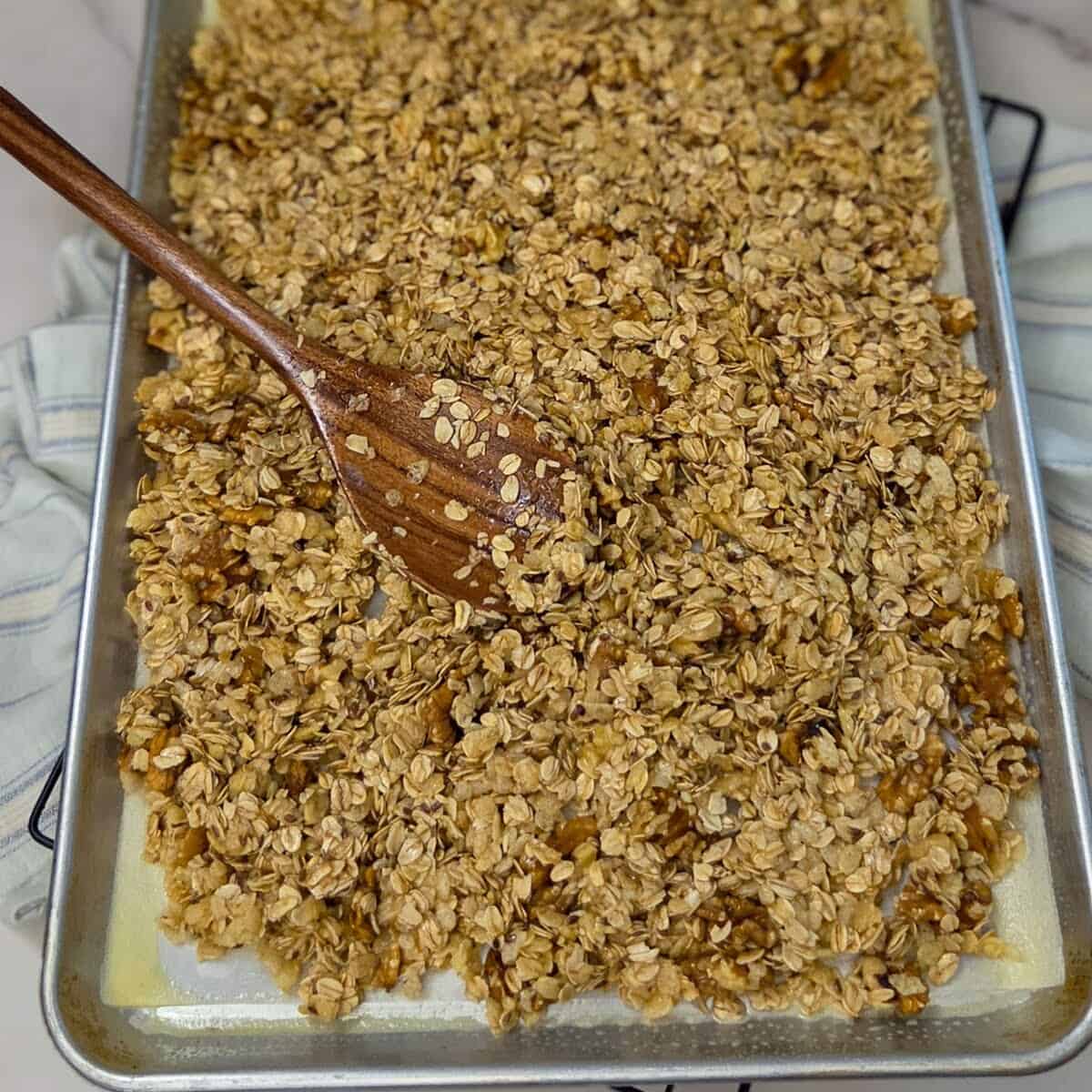 Spreading the banana walnut granola mixture onto a baking sheet pan lined with parchment paper with a wooden spoon.